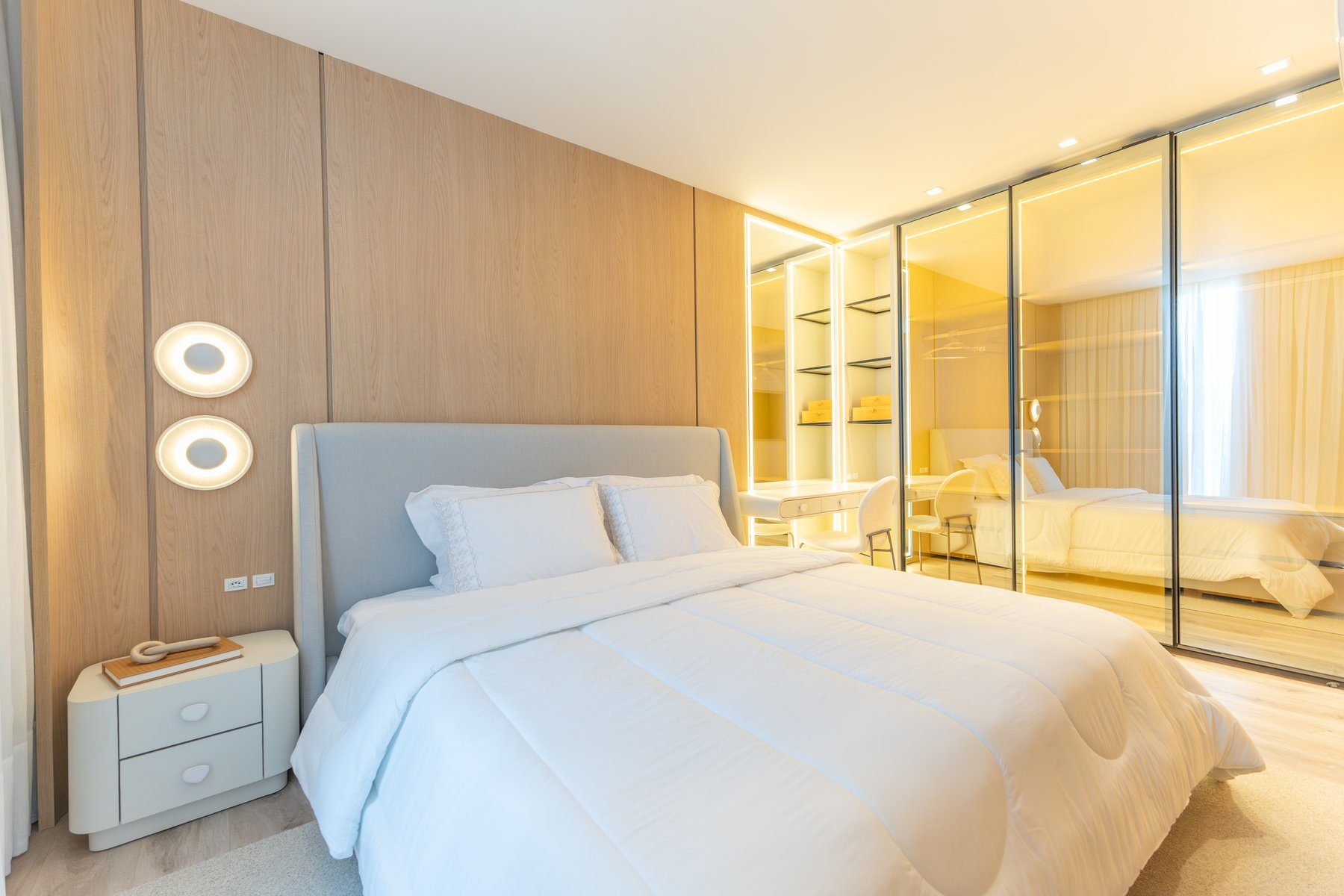 Quarto com cabeceira estofada, marcenaria em madeira clara e closet espelhado com iluminação indireta